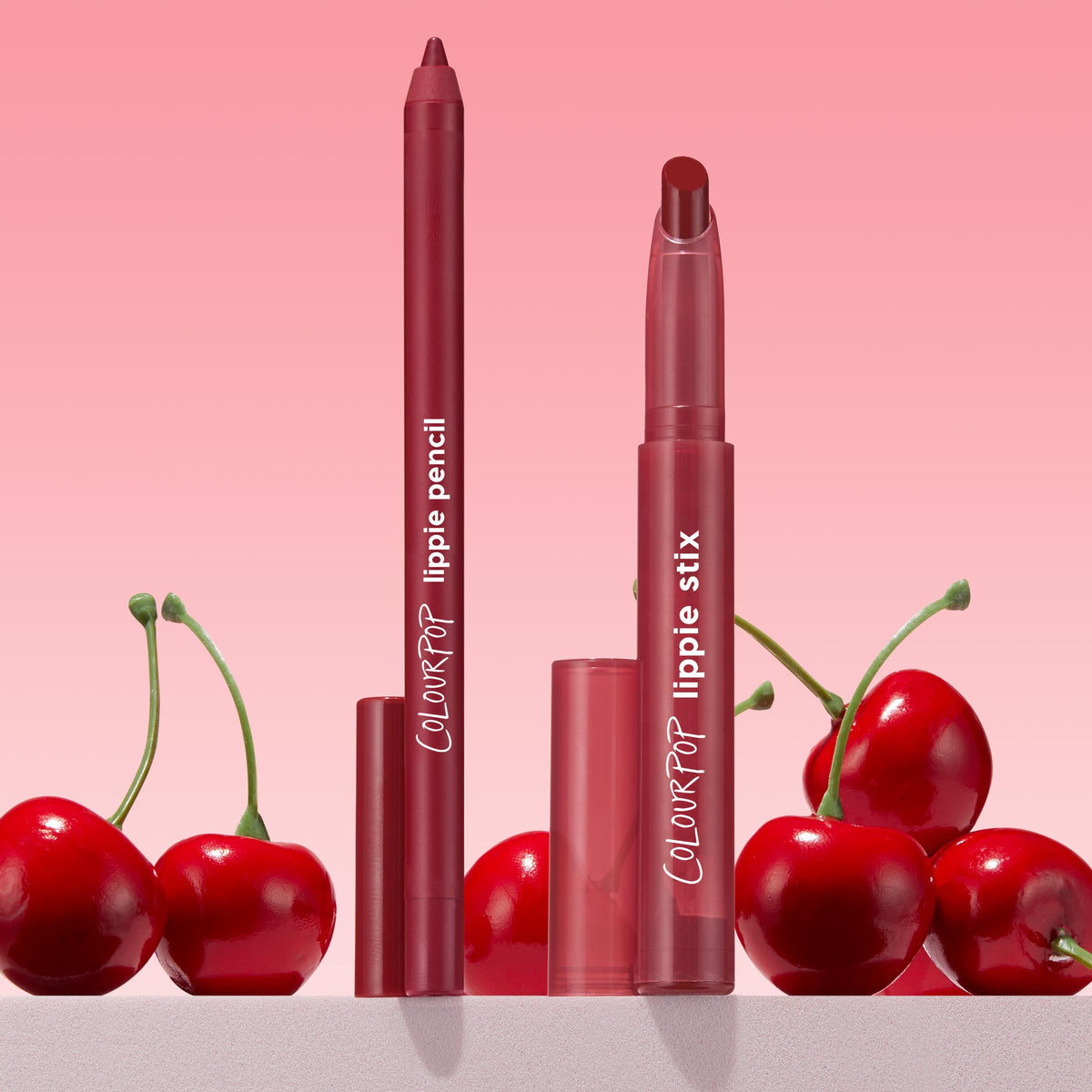 Bichette ColourPop Lippie Pencil and matching Lippie Stix stand upright with caps off, revealing rich cherry-red tips, surrounded by glossy cherries on a pink gradient backdrop, highlighting a creamy, precise liner duo for bold red lips.