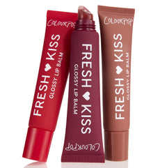 ColourPop Glossy Lip Balm Trio Get Rich tubes stand slightly angled, center tube uncapped to reveal a slanted applicator, in red, deep plum, and warm nude shades with Fresh Kiss branding on matte squeeze tubes against a white background.