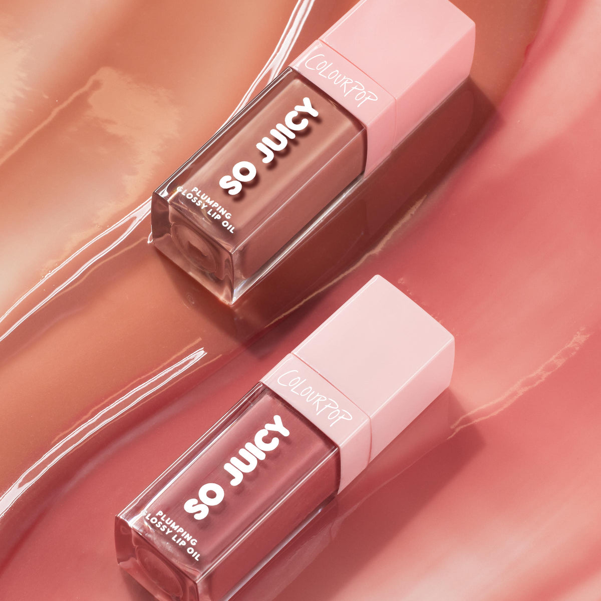 Two tubes of Juicy Moment, the ColourPop So Juicy Lip Oil Set, lie diagonally catching light; clear rectangular bottles with pink caps and bold SO JUICY lettering display plumping glossy lip oil against a swirled rose gloss backdrop.