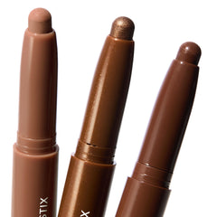 Three ColourPop Shadow Stix Essential Neutrals stand uncapped, revealing twist-up cream eyeshadow tips in nude beige, metallic bronze shimmer, and deep chocolate brown, arranged close-up on a clean white background.