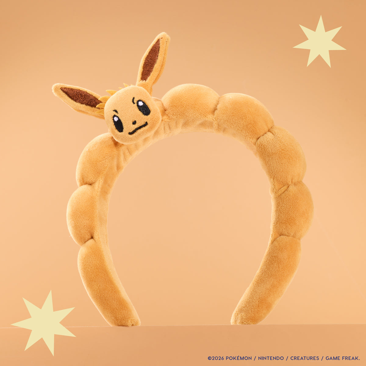 Plush Eevee Headband with a 3D Eevee head and ears on a padded tan band, peach backdrop with stars