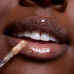 Close-up of glossy, glittering brown lips as a wand applies Ambitious Tiana Ultra Glossy Lip, a shimmery nude Disney Princess lip gloss, highlighting high-shine sparkle and smooth finish on deep skin tone.