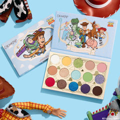 ColourPop The Toys Are Here shadow palette lays open, displaying 15 matte and shimmer shades - white, yellow, greens, blues, purple, pink, red, and neutrals - beside Toy Story character cover art, styled top-down with dolls and a cowboy hat on a sky-blue backdrop.