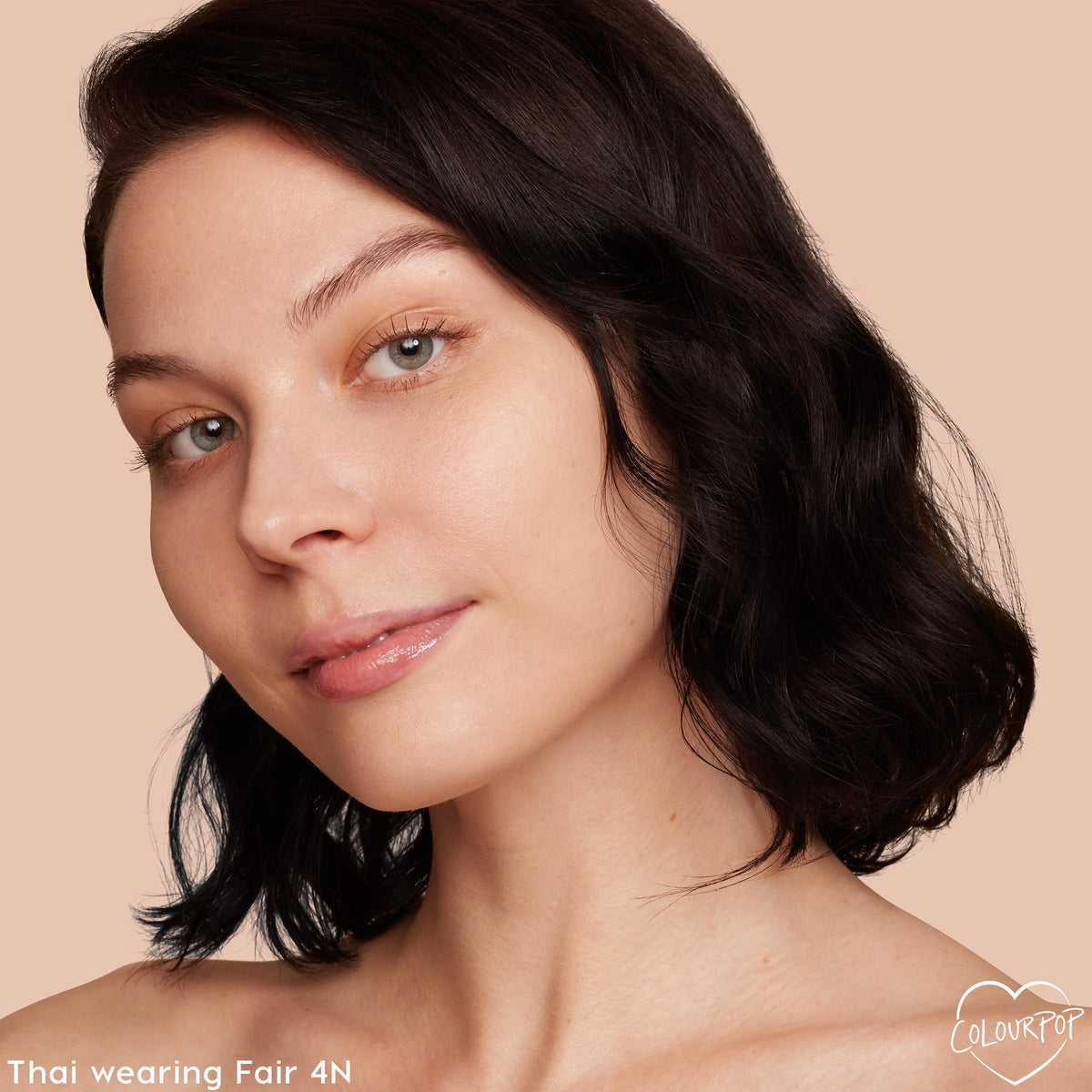 Model’s face wearing ColourPop Pretty Fresh Tinted Moisturizer in Fair 4 N, showcasing light dewy coverage and even skin tone; close-up three-quarter view with dark wavy hair and glossy lips against a soft peach background.