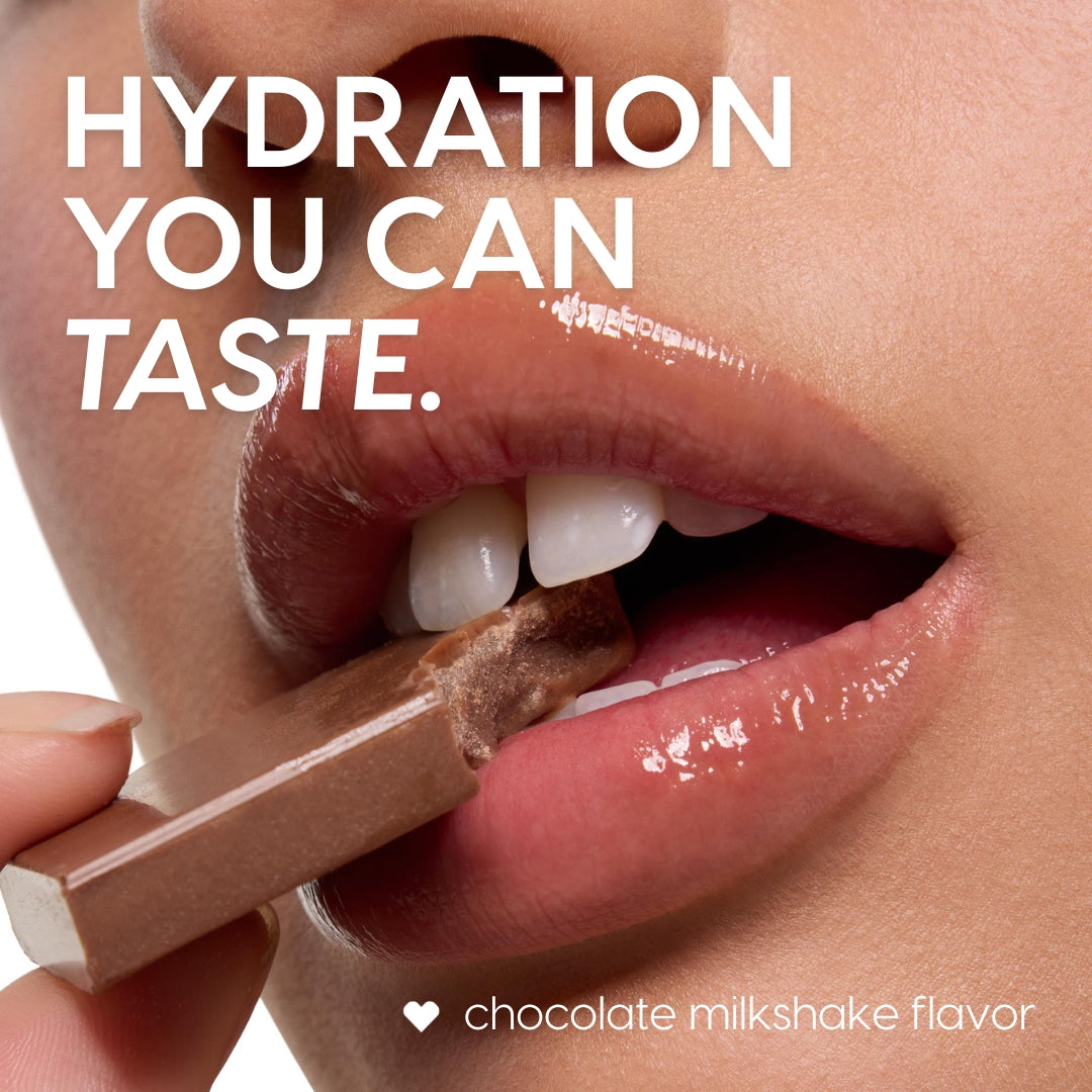 Glossy lips wear the Chocolate Milkshake ColourPop Lip Mask, biting a piece of milk chocolate to suggest flavor, with hydration shine visible in a tight beauty close-up, promoting a chocolate milkshake-flavored lip treatment.