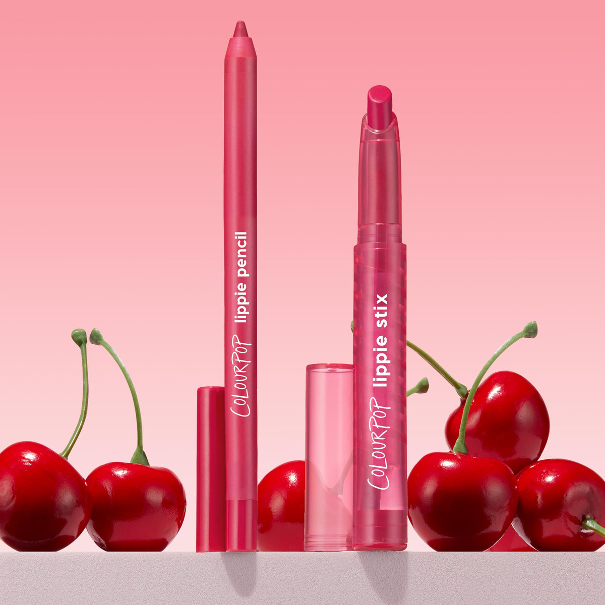 Ultimate Lip Combo showcases a pink-red ColourPop Lippie Stix + Pencil Set: lippie pencil and lippie stix standing upright with translucent pink caps, framed by fresh cherries against a soft pink gradient backdrop.