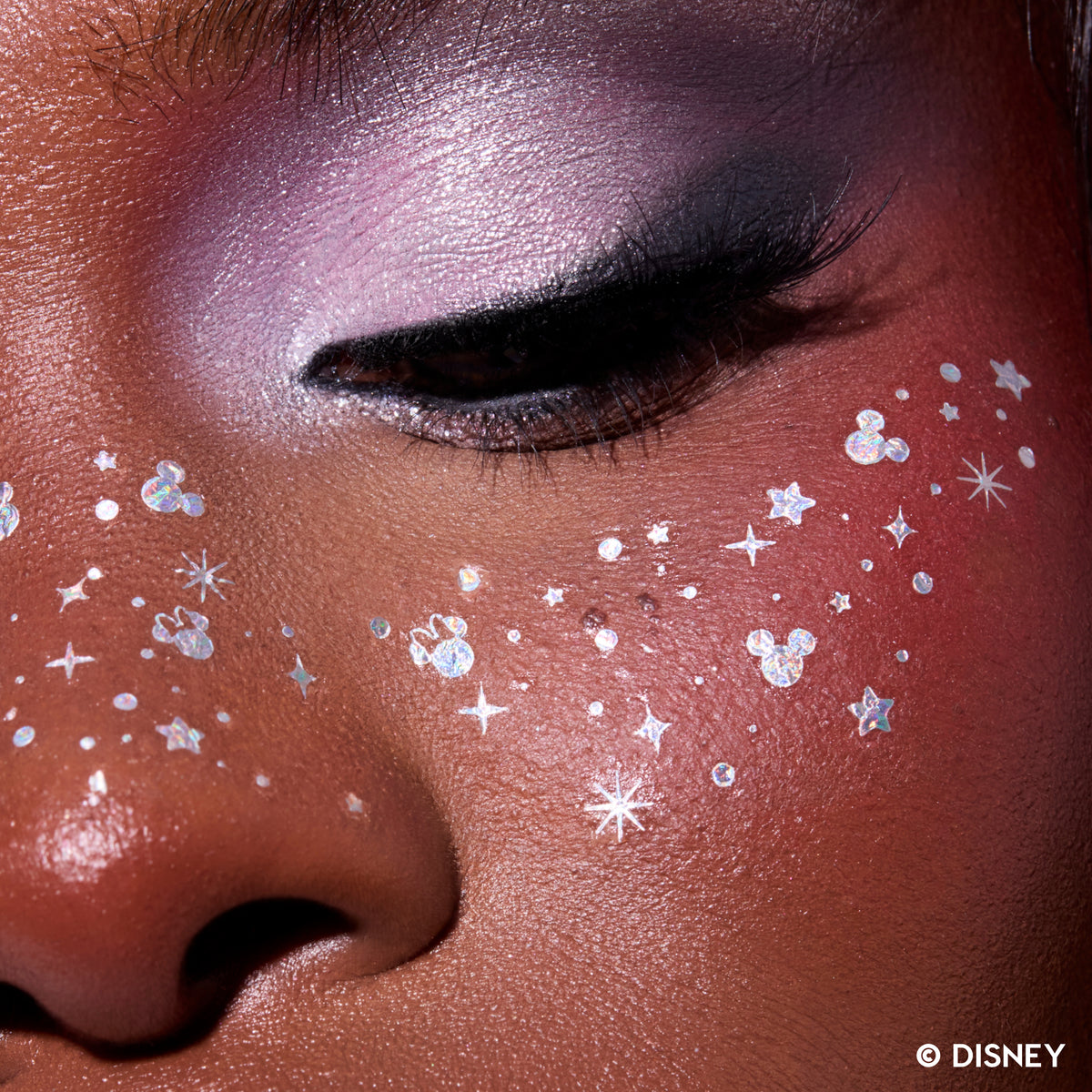 Close-up of glitter freckle tattoos with stars and tiny Mickey Mouse silhouettes across a deep-skin cheek and nose
