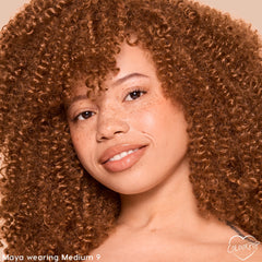 Model wearing ColourPop Pretty Fresh Tinted Foundation Balm in Medium 9, smiling in a warm studio close-up; dewy, natural coverage lets freckles show through, with glossy nude lips and tight curls framing the face.