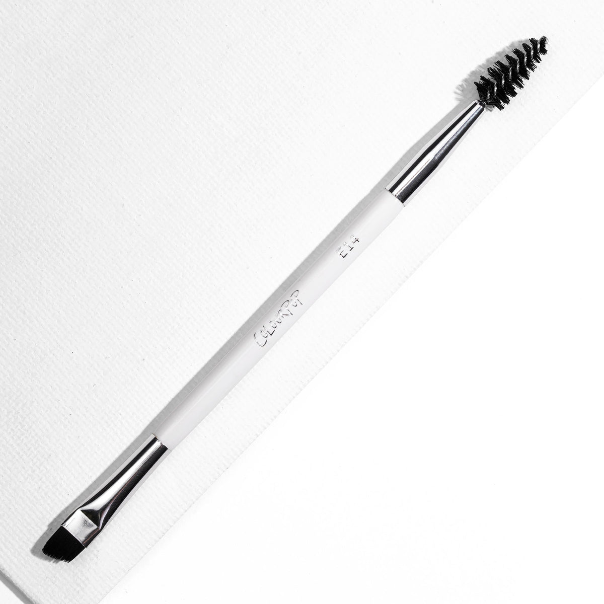 ColourPop Brow Dual Ended makeup brush resting diagonally, featuring an angled flat brow brush for filling and a black spoolie for grooming, white handle with chrome ferrules, on a bright white textured background.