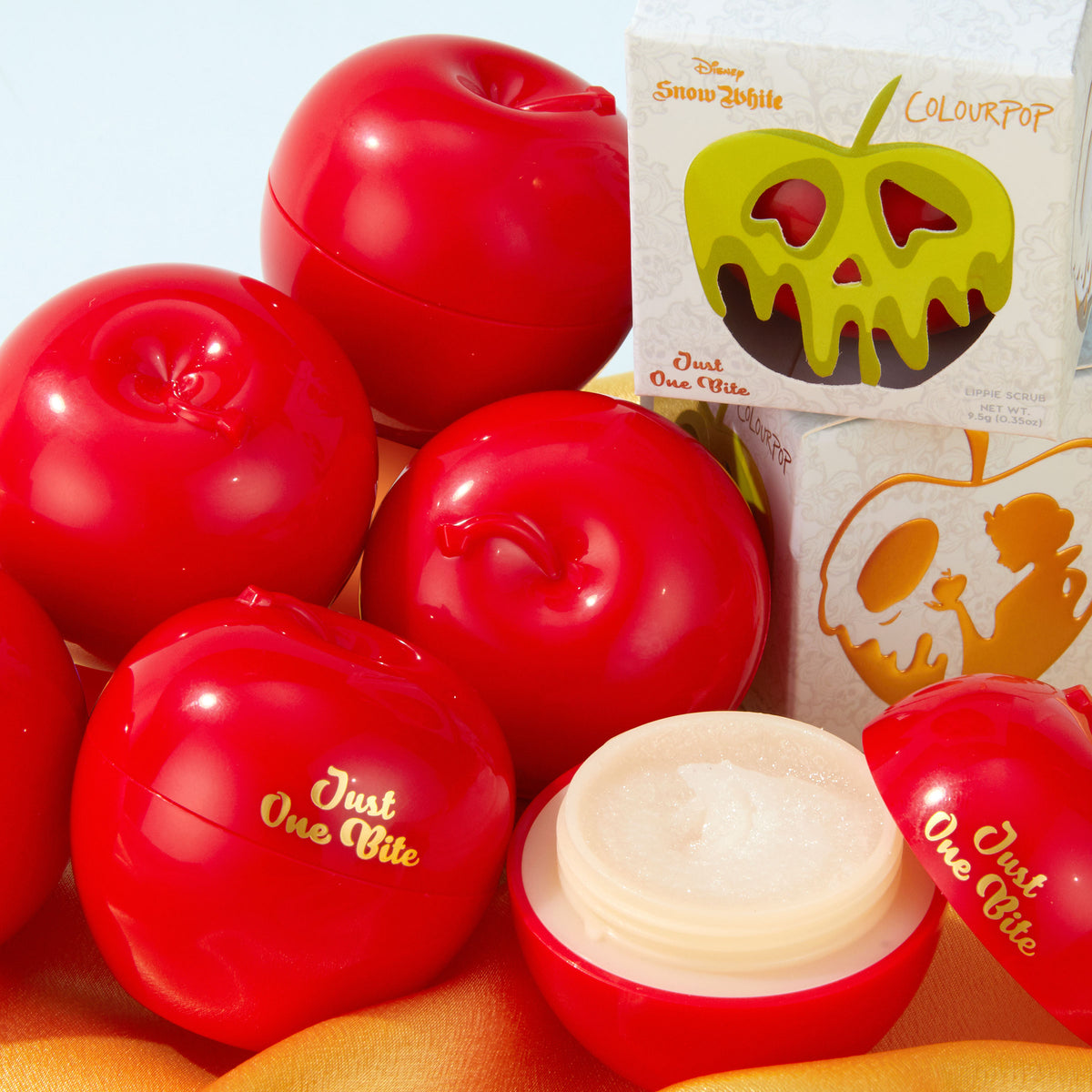 Just One Bite ColourPop Lip Scrub opens to reveal a sugary exfoliant in a glossy red apple-shaped jar, surrounded by matching apple containers and Disney Snow White packaging with a green poison-apple graphic; 0.3 oz lip scrub.