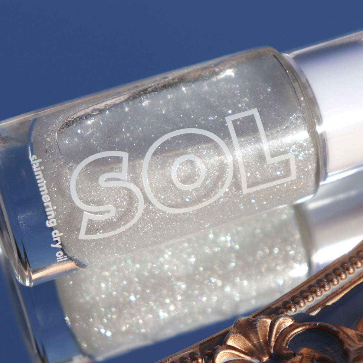 Glass bottle of ColourPop SOL Shimmering Dry Oil in Sterling Silver glistens with fine silver sparkle, resting angled against an ornate gold mirror frame under bright sunlight on a clear blue background.