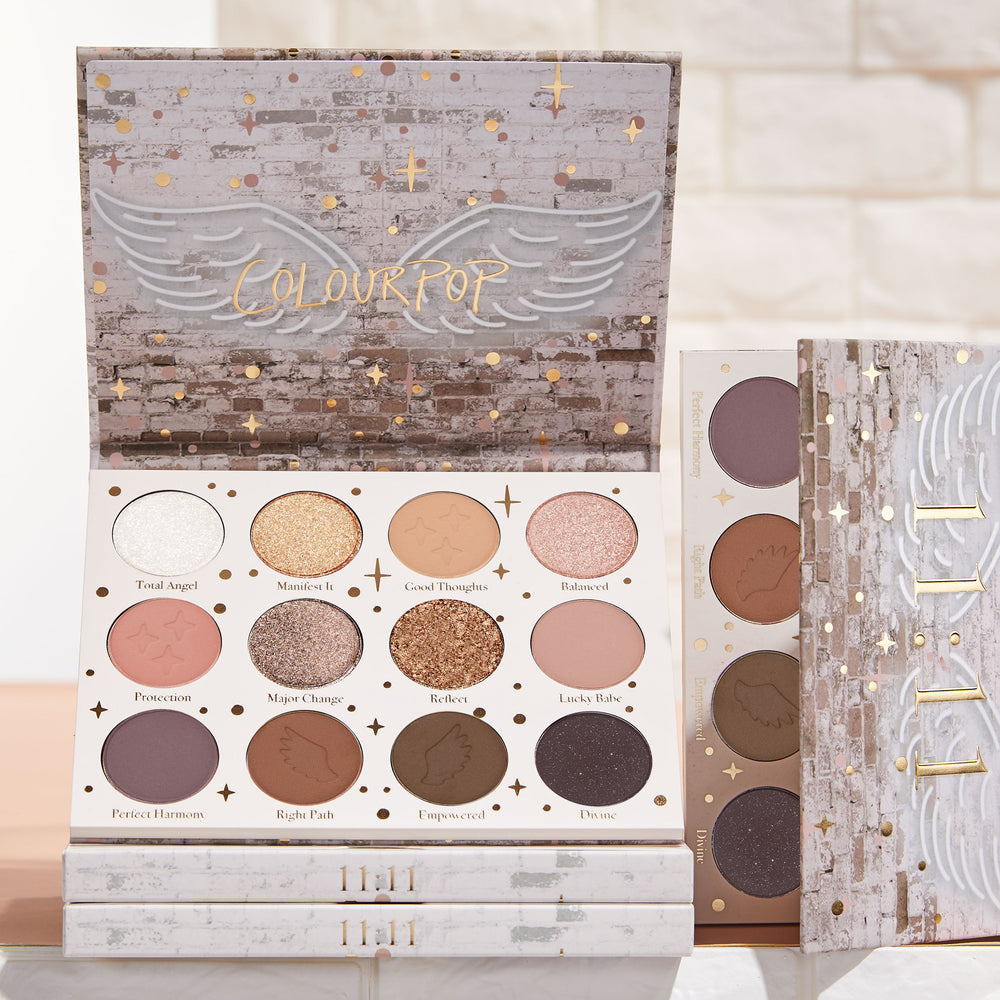 11:11 ColourPop Shadow Palette opens to 12 neutral and rosy tones - matte, shimmer, and pressed glitter - with star and wing embossing; displayed in brick-and-gold celestial packaging with stacked palettes beside it in soft studio light.