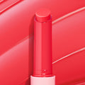 Delulu shade of the ColourPop So Juicy Plumping Gloss Balm with Peptides, twist-up bullet extended upright from a sleek tube with a light pink base, glossy coral-red stick centered against a swirled pink backdrop.