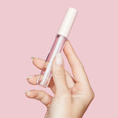 ColourPop Ultra Glossy Lip 20 Carats tube held between fingers, showing clear gloss in a transparent bottle with a pale pink cap and white logo, against a pastel pink background.