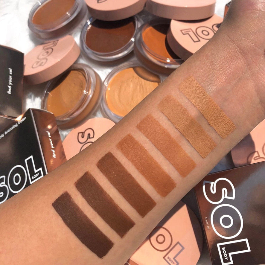 Fair shade of ColourPop SOL Face & Body Bronzing Balm swatched on a forearm beside deeper bronzer stripes, showing smooth coverage; round SOL Body compacts with blush-pink lids and boxes scattered in the background.