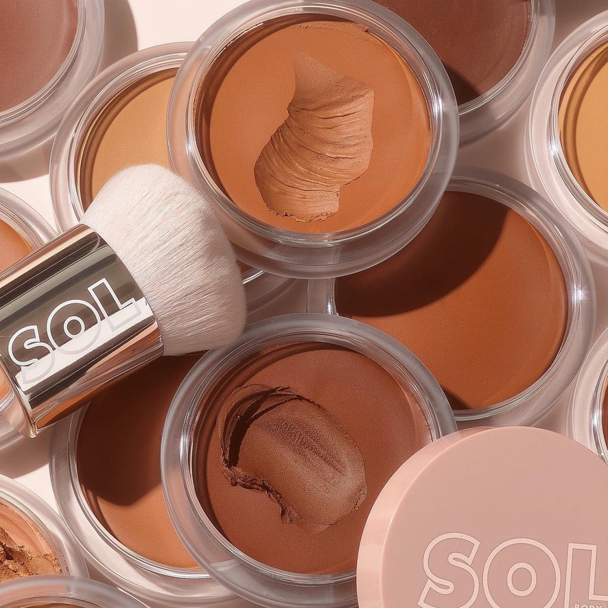 Deep shade of ColourPop SOL Face & Body Bronzing Balm swatched in an open clear pot, surrounded by other warm tones and a SOL-branded kabuki brush in soft light, showcasing the creamy texture for sun-kissed face and body.