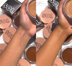 Medium Dark ColourPop SOL Face & Body Bronzing Balm - arm swatch shows before-and-after glow, left bare wrist vs right sun-kissed tan; round SOL pots and boxes surround the hand holding an open balm under soft indoor light.