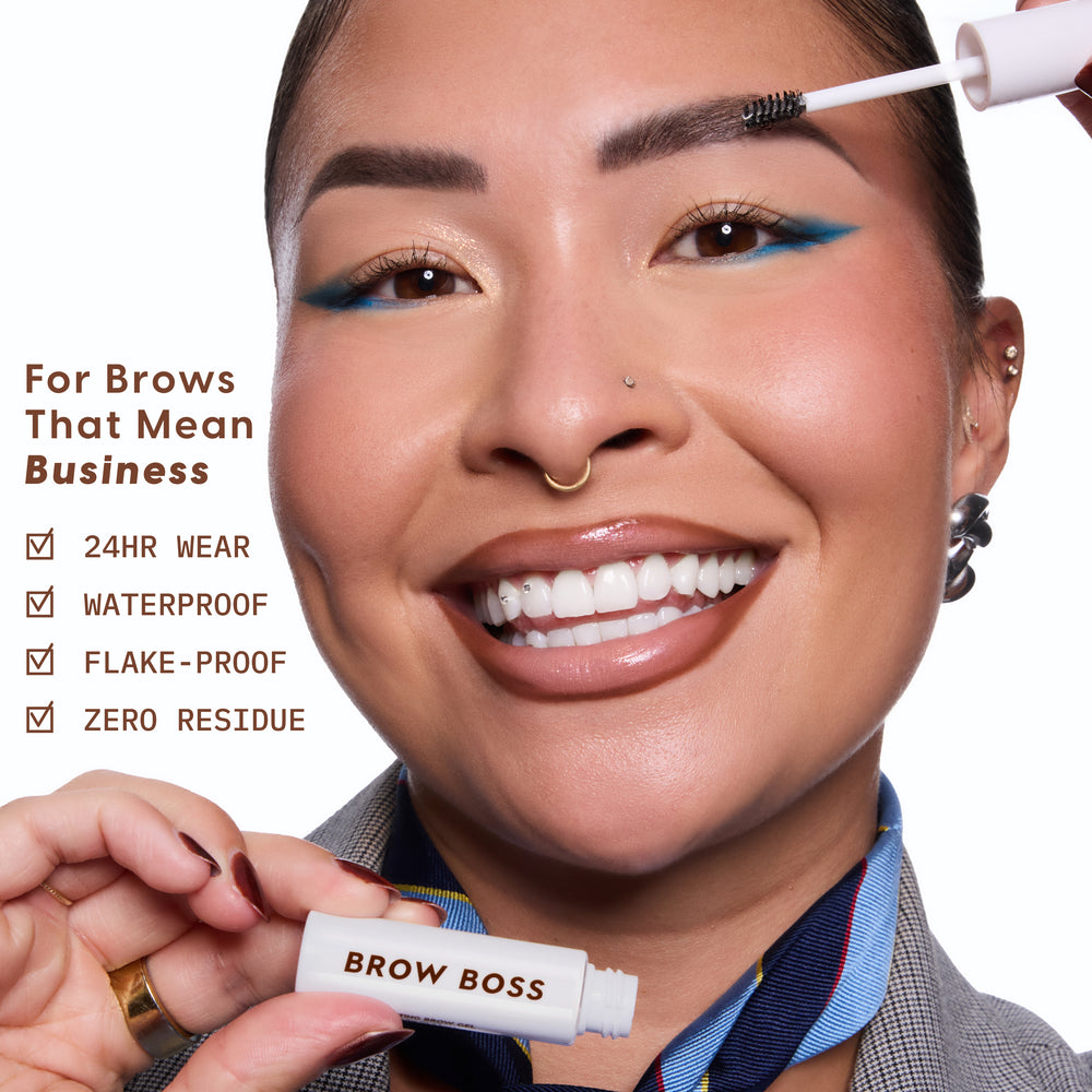 Smiling model brushes Clear Brow Boss Gel onto groomed eyebrows, showcasing bold blue eyeliner, while holding the white Brow Boss tube beside text highlighting 24HR wear, waterproof, flake-proof, zero residue brow makeup benefits.