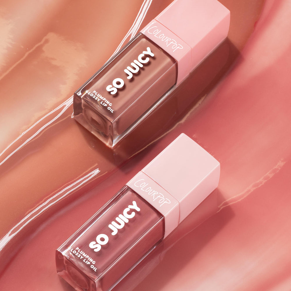 Two tubes of Juicy Moment, the ColourPop So Juicy Lip Oil Set, lie diagonally catching light; clear rectangular bottles with pink caps and bold SO JUICY lettering display plumping glossy lip oil against a swirled rose gloss backdrop.