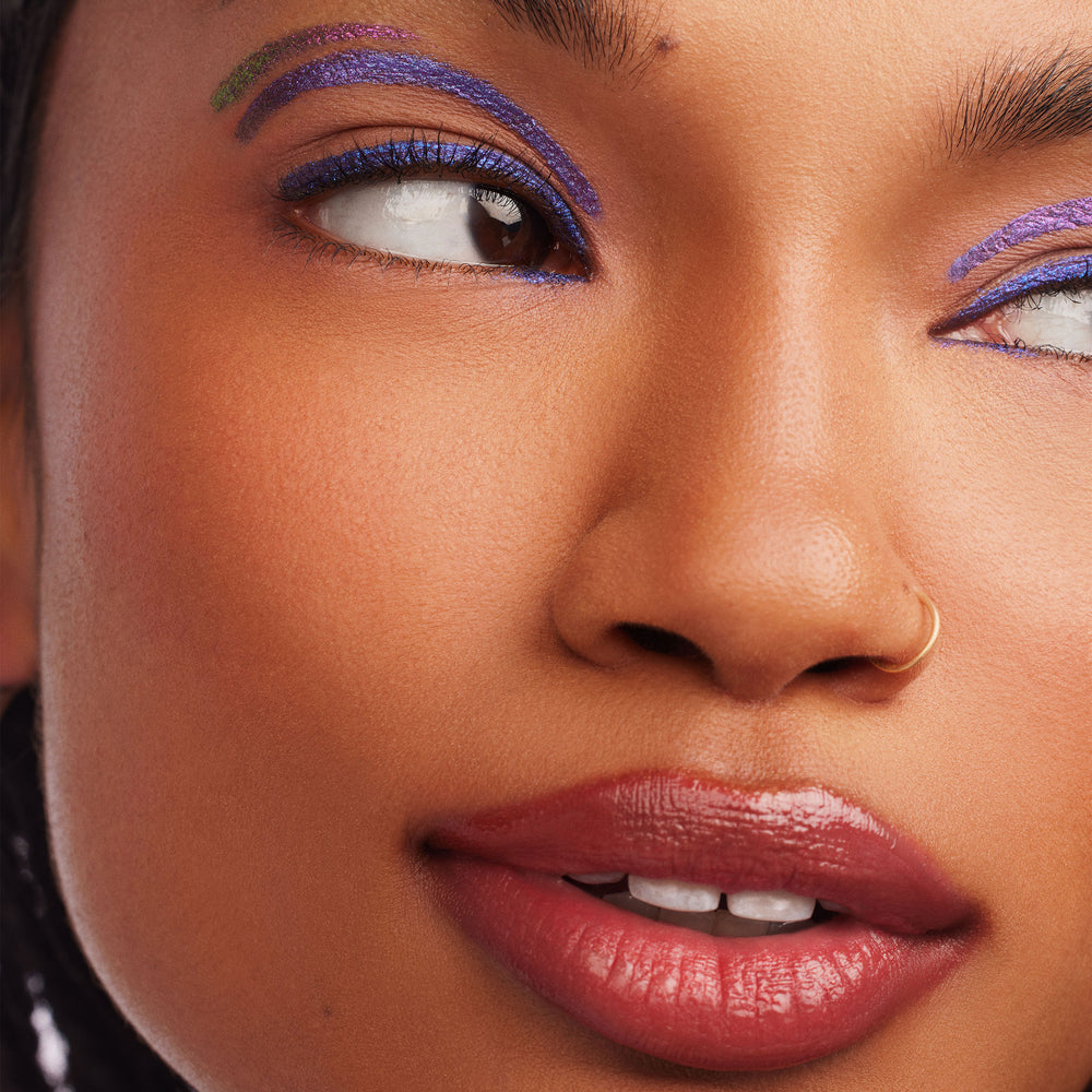 Supernova ColourPop Chrome Liquid Shadow shimmering multi‑chrome purple with a green shift, arcing above the crease and lining both eyes in a bold graphic look, shown in a close-up with glossy lips and a small gold nose ring.