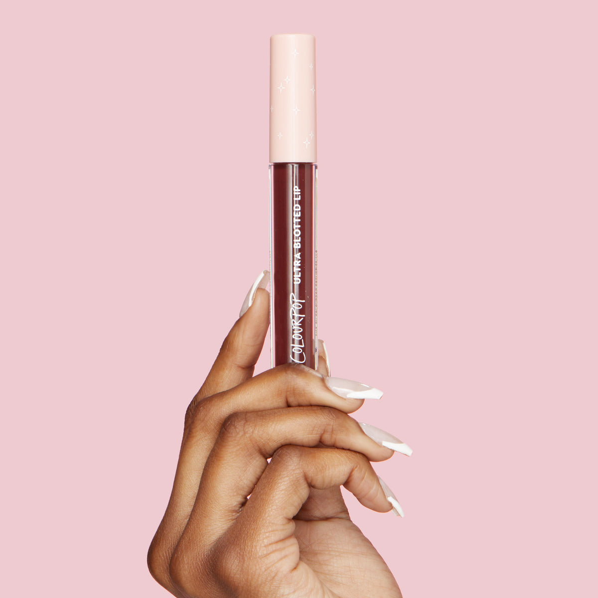 ColourPop Ultra Blotted Lip in Canon Dr held upright by a manicured hand, showcasing a clear tube of deep berry lip color with a pale pink star-print cap and white logo, set against a solid pink backdrop.
