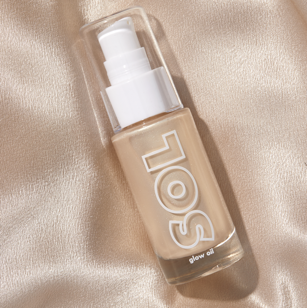 Clear glass pump bottle of Champagne SOL Glow Oil resting diagonally on shimmering champagne satin, the pearlescent golden formula visible behind the SOL logo; ColourPop SOL Glow Oil body highlighter for a luminous glow.