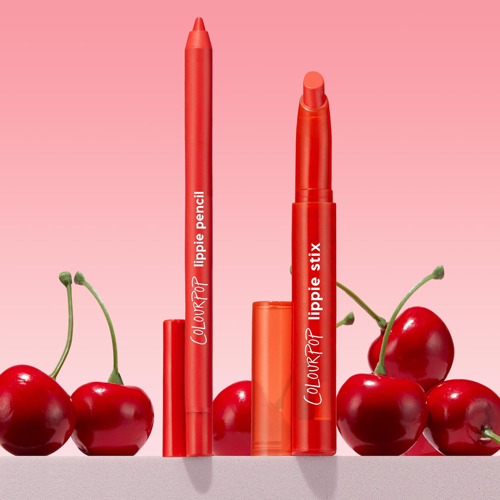 Sealed with a Kiss ColourPop Lippie Stix + Pencil Set stands uncapped, showing a creamy cherry-red Lippie Stix and matching lip pencil, posed amid fresh cherries on a light surface against a pink gradient background.