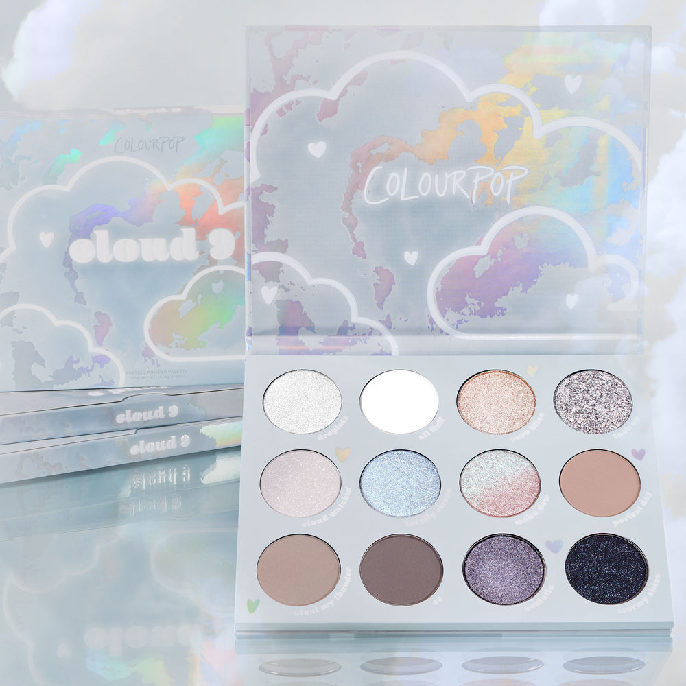 Cloud 9 ColourPop Shadow Palette opens to reveal 12 cool-toned neutrals and iridescent shimmers - from icy silver and matte white to taupe, lilac, and deep navy - set in holographic cloud packaging with stacked palettes and pastel clouds behind.