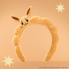 Plush Eevee Headband with a 3D Eevee head and ears on a padded tan band, peach backdrop with stars