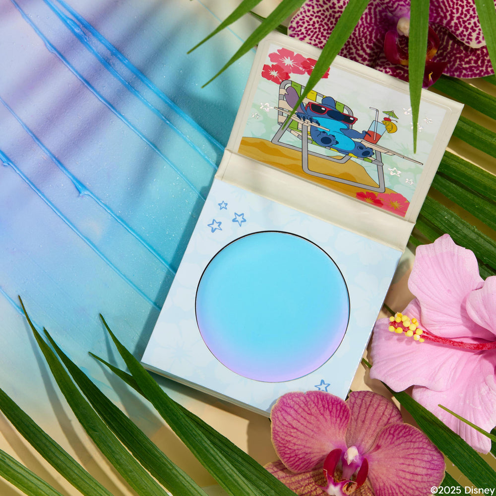 Crowd Favorite ColourPop Cream Blush compact sits open, revealing a round pan and inner lid art of Stitch lounging on a beach chair, set in a tropical flat lay with palm fronds, pink orchids, hibiscus, and a blue gradient backdrop.