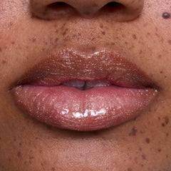 Close-up of glossy, medium-toned lips wearing Ambitious Tiana Ultra Glossy Lip, showing shimmering gold and pink sparkle inspired by Disney Princess beauty, with natural skin and freckles softly visible around the mouth.