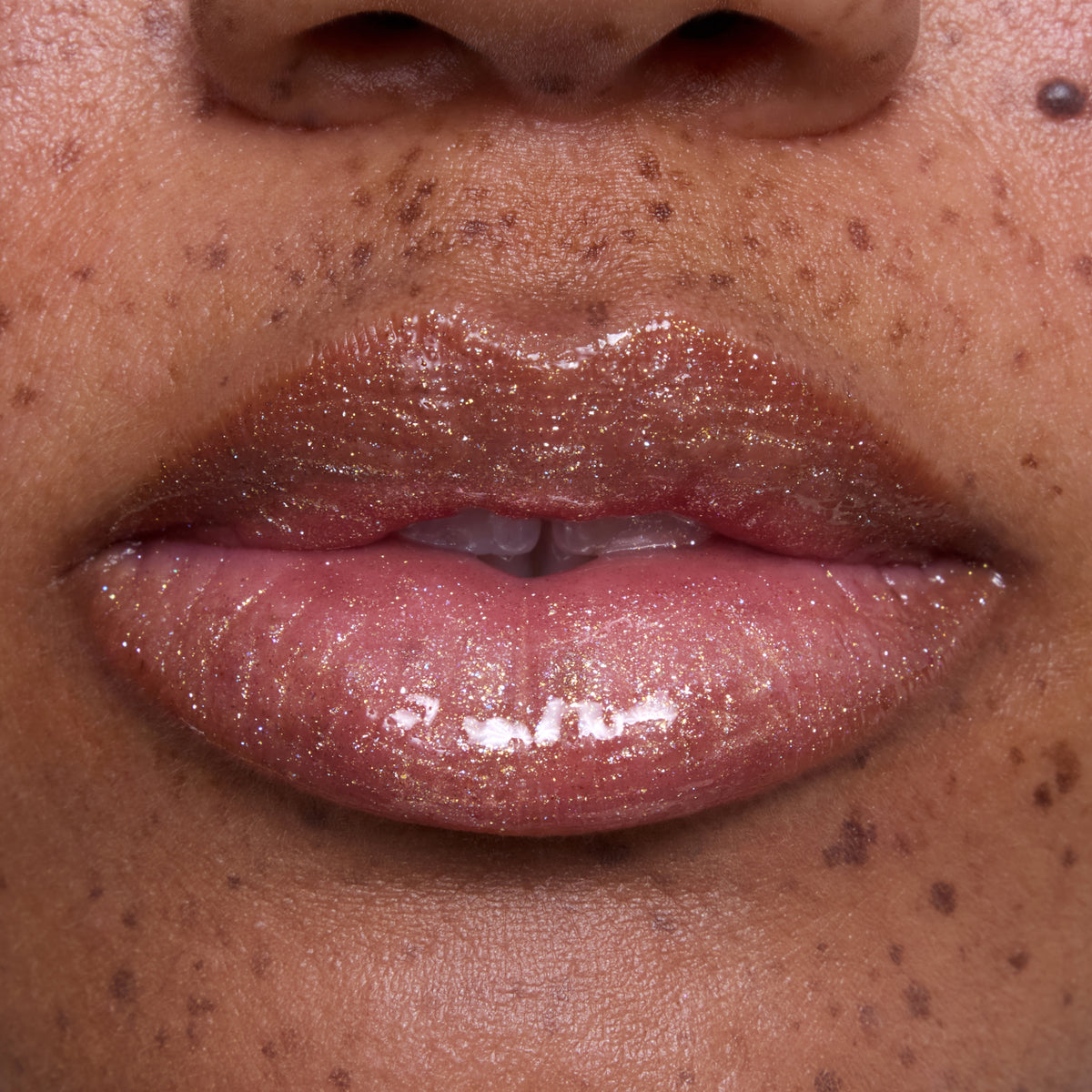Close-up of glossy, medium-toned lips wearing Ambitious Tiana Ultra Glossy Lip, showing shimmering gold and pink sparkle inspired by Disney Princess beauty, with natural skin and freckles softly visible around the mouth.
