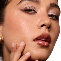 Glossed lips wearing ColourPop So Juicy Plumping Gloss Balm with Peptides, shade Perfection, delivering a plumped high-shine finish as a model touches her cheek; close-up beauty shot with neutral eyeshadow in studio lighting.