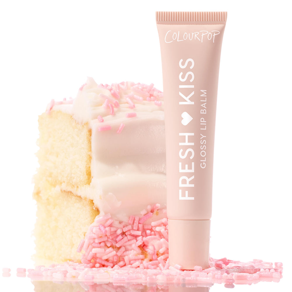 ColourPop Fresh Kiss Glossy Balm, Vanilla Cake, stands upright against a frosted vanilla cake slice topped with pink sprinkles, blush tube with white lettering styled on a bright, reflective surface for a sweet, high-shine lip finish.