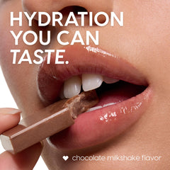 Glossy lips wear the Chocolate Milkshake ColourPop Lip Mask, biting a piece of milk chocolate to suggest flavor, with hydration shine visible in a tight beauty close-up, promoting a chocolate milkshake-flavored lip treatment.