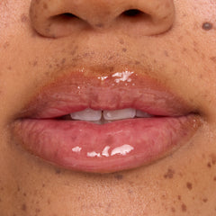 Glossy, hydrated lips wear a clear lip mask with small white balm flecks melting on the surface, shown in an extreme close-up on freckled skin to showcase ColourPop lip mask treatment shine and texture.