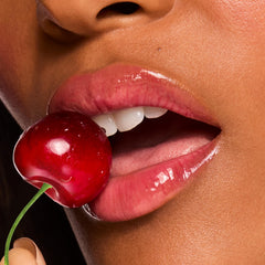 Glossy pink lips wear ColourPop Fresh Kiss Glossy Balm in Cherry Glaze, gripping a red cherry between the teeth in a macro beauty shot, showcasing a high-shine, juicy finish and hydrating lip balm texture.