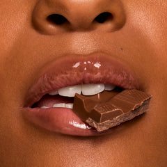 Glossy, cocoa-tinted lips wearing ColourPop Fresh Kiss Glossy Balm in Chocolate Milkshake hold a square of milk chocolate between teeth in a macro close-up, showcasing high-shine, cushy hydration and a creamy nude tint.