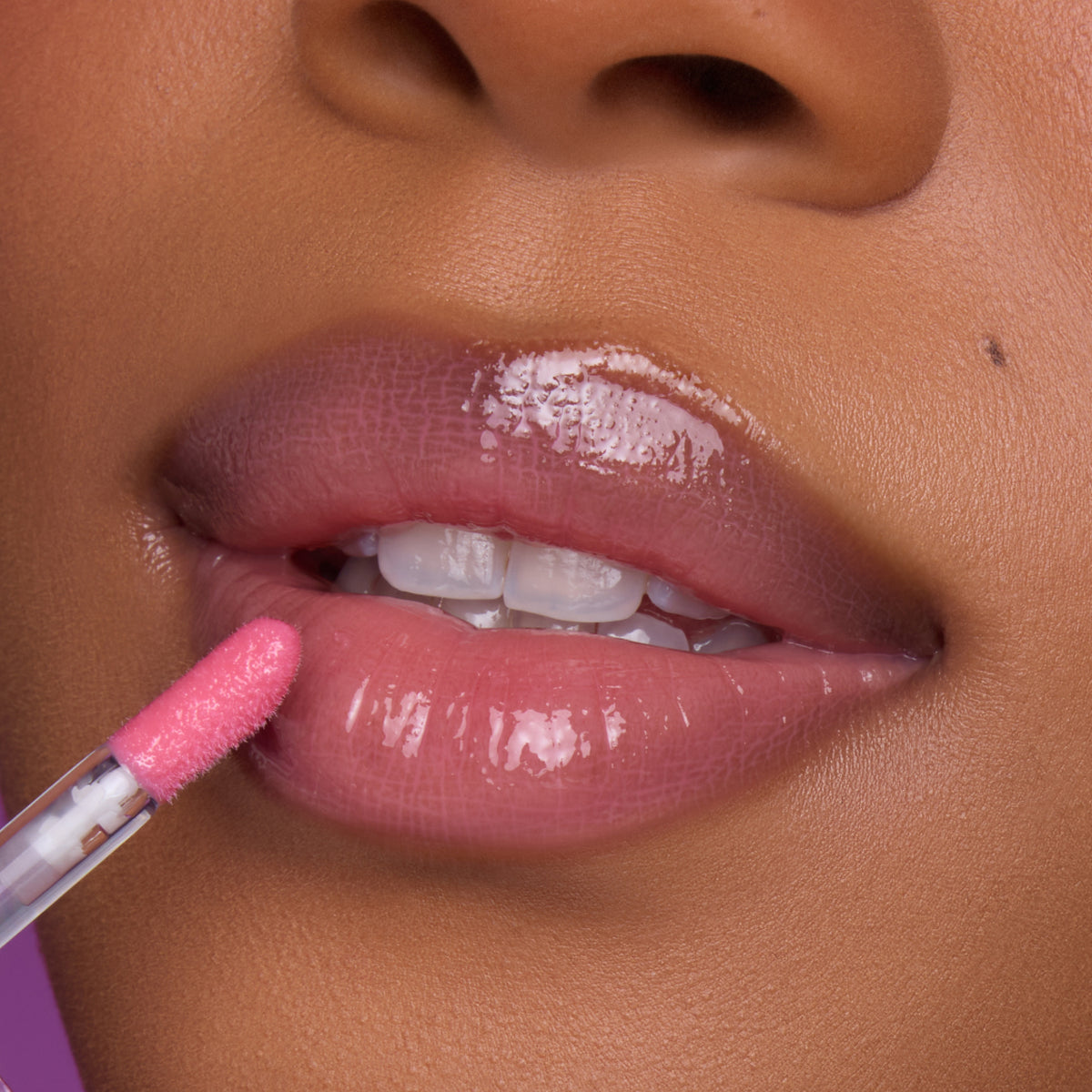 Close-up of a model’s lips applying Still That Girl Ultra Glossy Lip from Heart to Get, showing a sheer pink, high-shine gloss on smooth lips against a soft purple background for a glamorous beauty look.