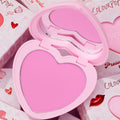 Heart-shaped Hot 2 Touch compact opens to reveal bright pink ColourPop Pressed Powder Blush, set in a pastel pink case with built-in mirror, resting atop Valentine-themed boxes with hearts and red lip graphics.