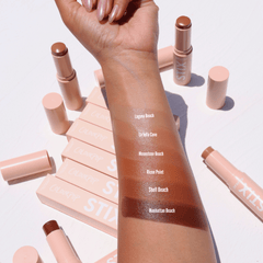 ColourPop Bronzer Stix in Laguna Beach swatched on a forearm - a warm, sun-kissed tan stripe - with additional shade swatches below and nude-pink STIX tubes and boxes scattered on a white surface in bright daylight.