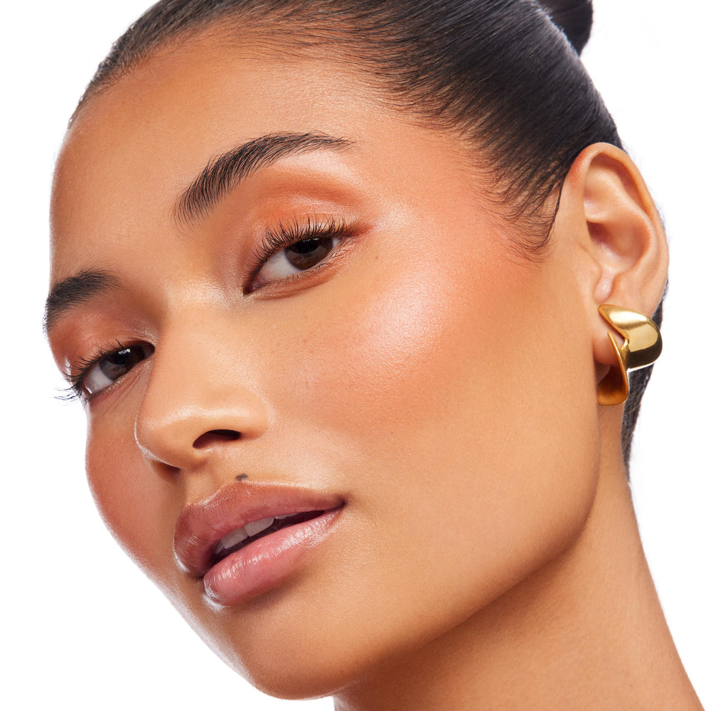 Molten Hot Lite Stix highlighter glowing on a model’s cheekbone, creating a warm peachy-gold sheen; ColourPop Lite Stix swept along cheek and brow bone in a studio close-up with soft coral eye makeup and a sculptural gold earring.