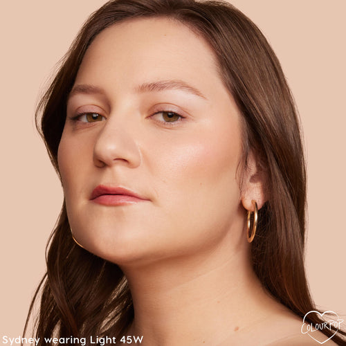 Model wearing Light 45 W, a ColourPop Hyaluronic Hydrating Foundation, showing smooth, dewy medium coverage with warm undertones; close-up of face angled left with natural brows and soft pink lip, gold hoop earring, on a peach background.