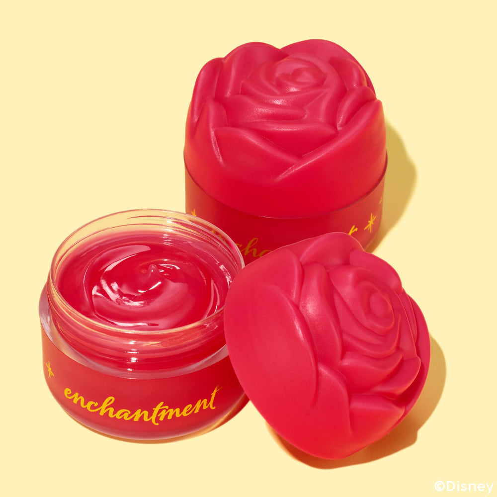 Enchanted Rose ColourPop Lip Mask shown open, revealing a swirled glossy raspberry-pink balm, flanked by two sculpted rose lids; red frosted glass jar with gold “enchantment” text on a pale yellow background.