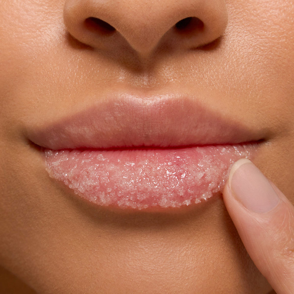 Strawberry Milk ColourPop Lip Scrub being applied as a fingertip massages pink sugar crystals across the lower lip in a tight close-up, showcasing glossy exfoliation and soft, hydrated lips.