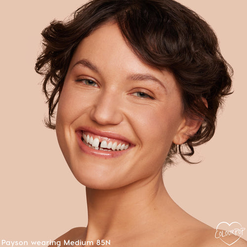 Model smiles in a studio close-up wearing ColourPop Hyaluronic Hydrating Foundation, shade Medium 85N, showcasing a hydrated, dewy medium coverage and even tone against a peach background.
