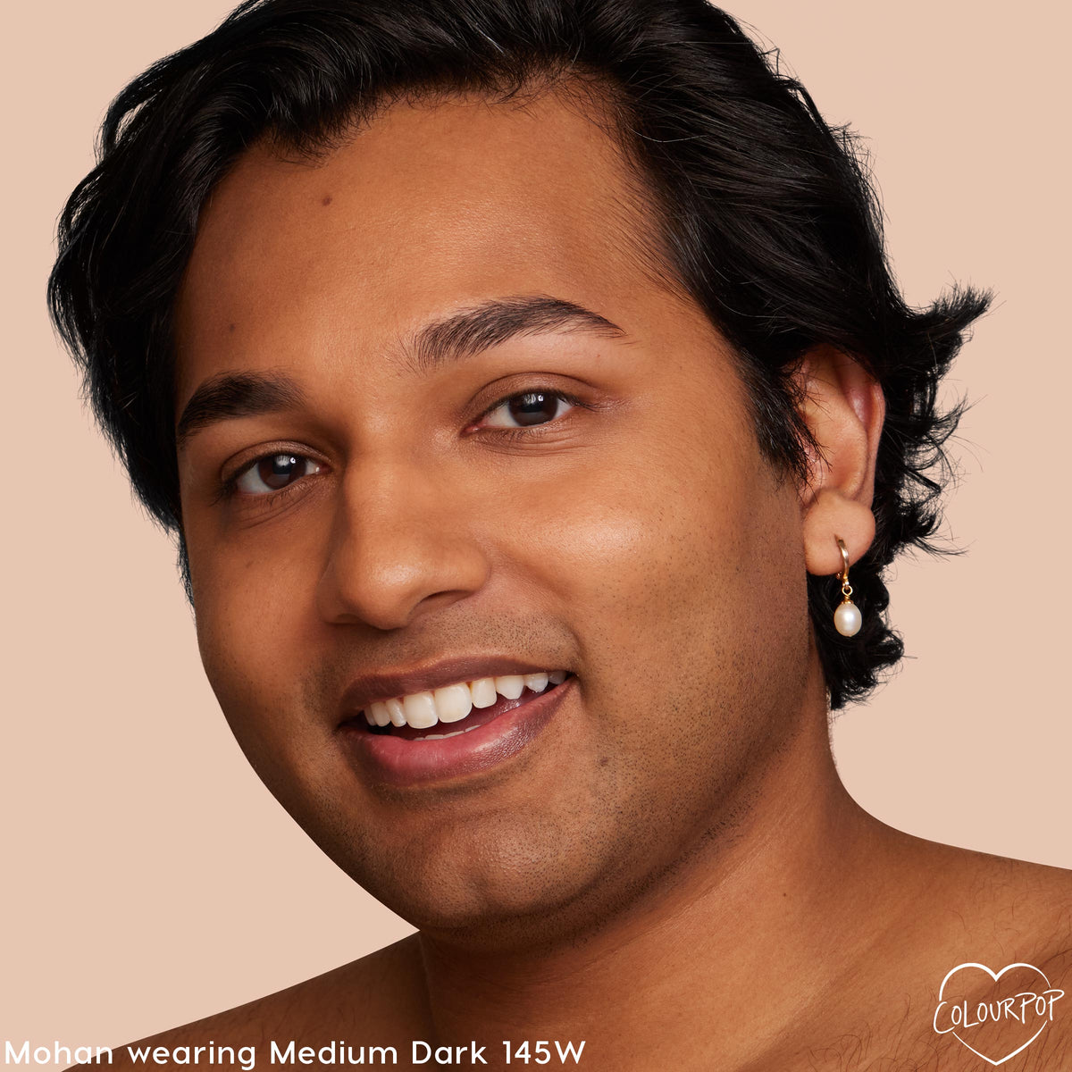 Model smiles wearing ColourPop Pretty Fresh Creamy Concealer in shade Medium Dark 145 W, showcasing smooth, natural coverage; close-up face on a peach-beige background with a gold hoop pearl earring.