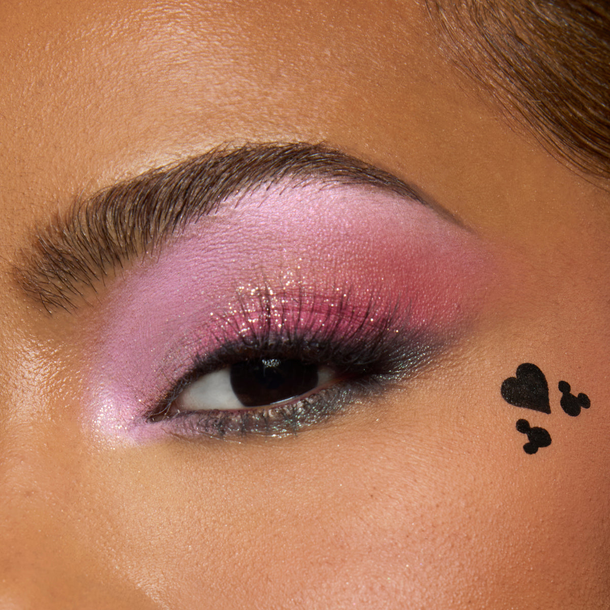 Close-up of a medium-skin eye with pink shimmer shadow and a black heart and Mickey Mouse stamp on the cheekbone