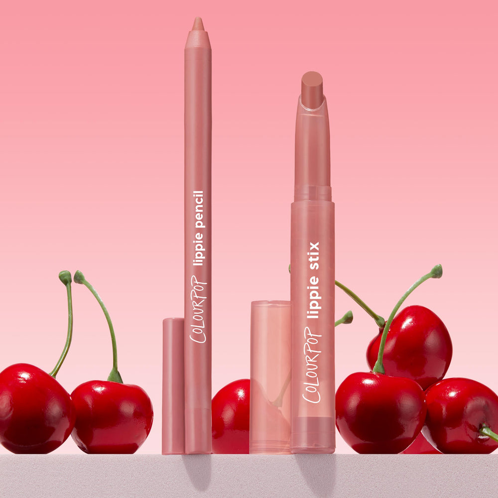 ColourPop Lippie Pencil "Oh Snap" stands uncapped with a creamy soft pink-nude tip, paired with matching Lippie Stix, on a countertop with glossy red cherries against a pink gradient backdrop - ideal for precise, long-wear lip lining.