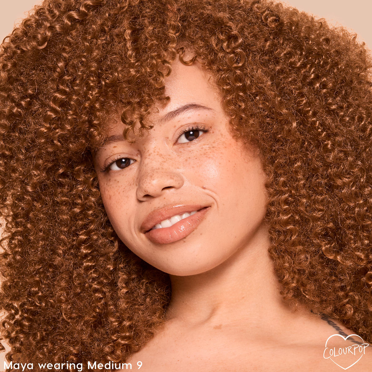 Model wearing ColourPop Pretty Fresh Tinted Foundation Balm in Medium 9, smiling in a warm studio close-up; dewy, natural coverage lets freckles show through, with glossy nude lips and tight curls framing the face.